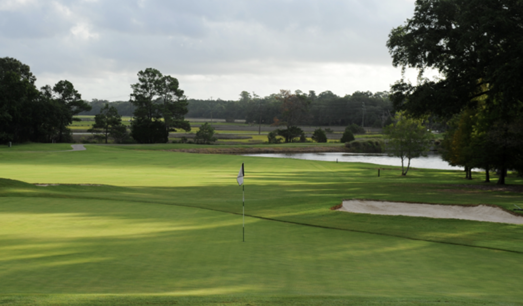Golf course green at Snee Farm Country Club in Mount Pleasant, SC, featuring a putting green, flagstick, sand bunker, pond, and mature Lowcountry landscaping.